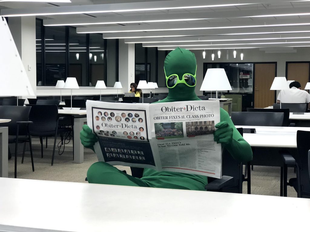 Humorous photo of a person in an alien costume reading Obiter in the library